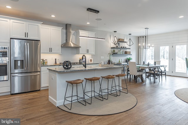 kitchen with white cabinets, stainless steel appliances, hardwood / wood-style floors, wall chimney range hood, and hanging light fixtures