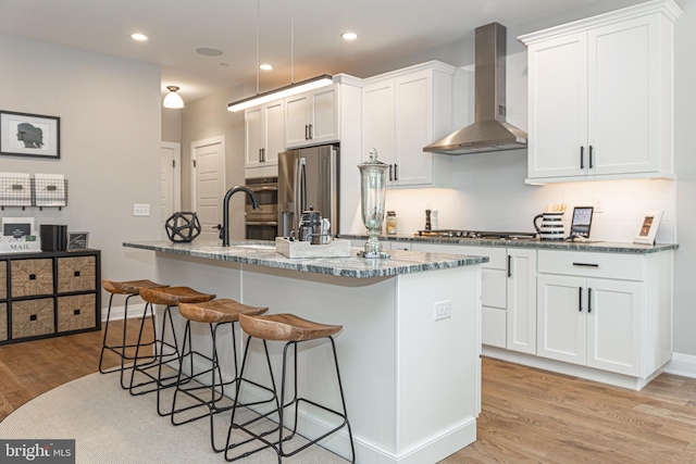 kitchen with appliances with stainless steel finishes, a breakfast bar, wall chimney exhaust hood, white cabinetry, and light wood-type flooring