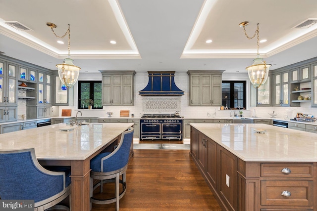 kitchen with light stone counters, custom exhaust hood, a tray ceiling, decorative light fixtures, and a large island