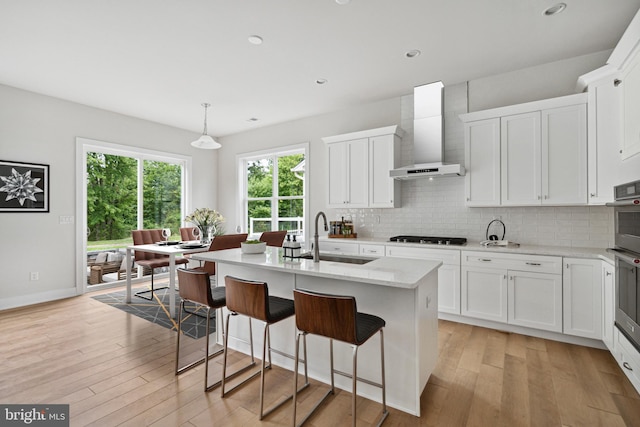 kitchen with tasteful backsplash, a center island with sink, white cabinetry, light hardwood / wood-style flooring, and wall chimney exhaust hood