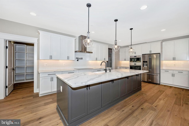 kitchen with wall chimney exhaust hood, stainless steel appliances, an island with sink, and white cabinets