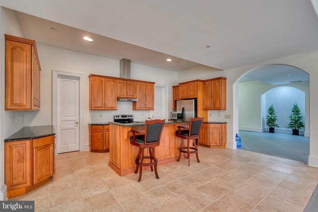 kitchen featuring a kitchen island with sink, appliances with stainless steel finishes, light tile floors, dark stone countertops, and a breakfast bar area