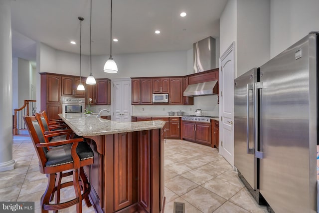 kitchen featuring hanging light fixtures, wall chimney range hood, stainless steel appliances, a kitchen breakfast bar, and a center island with sink