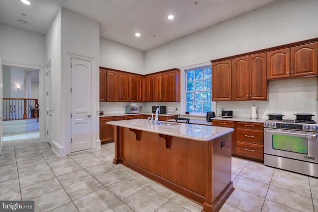 kitchen featuring light tile flooring, an island with sink, stainless steel range oven, sink, and light stone countertops