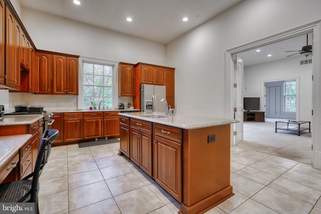 kitchen with an island with sink, light colored carpet, ceiling fan, light stone counters, and stainless steel fridge with ice dispenser