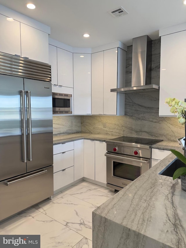 kitchen featuring wall chimney range hood, white cabinetry, light stone counters, and built in appliances