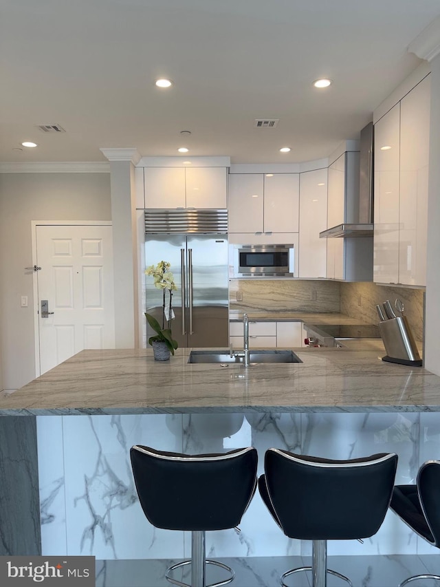 kitchen featuring white cabinetry, crown molding, built in appliances, kitchen peninsula, and wall chimney range hood