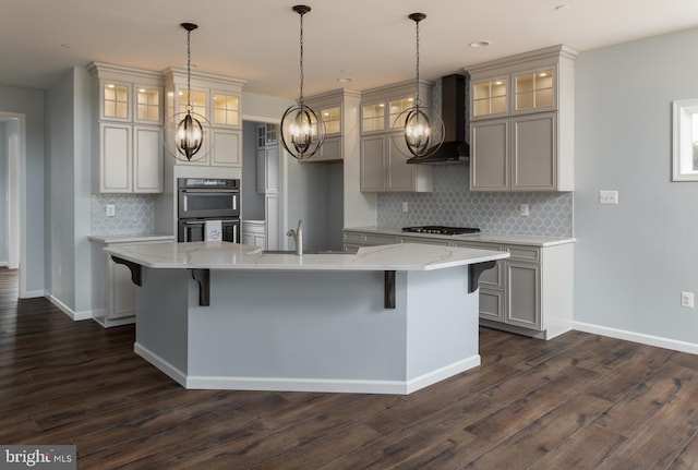 kitchen with black double oven, sink, an island with sink, and wall chimney range hood