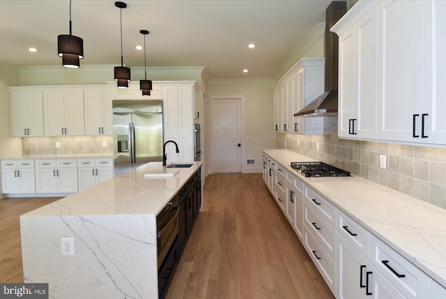 kitchen with wall chimney exhaust hood, stainless steel appliances, light hardwood / wood-style flooring, decorative light fixtures, and a center island with sink