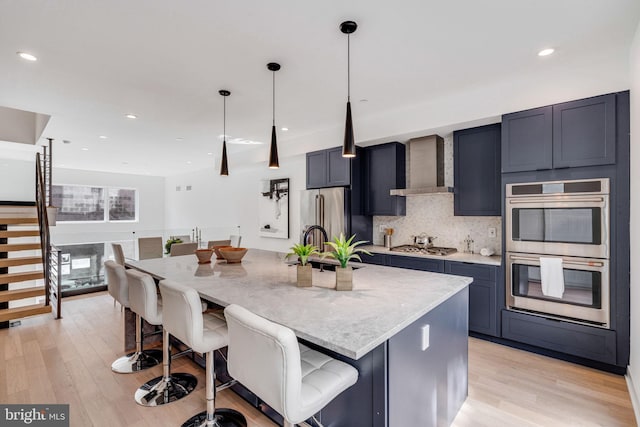 kitchen with stainless steel appliances, wall chimney range hood, light hardwood / wood-style flooring, decorative light fixtures, and a center island with sink