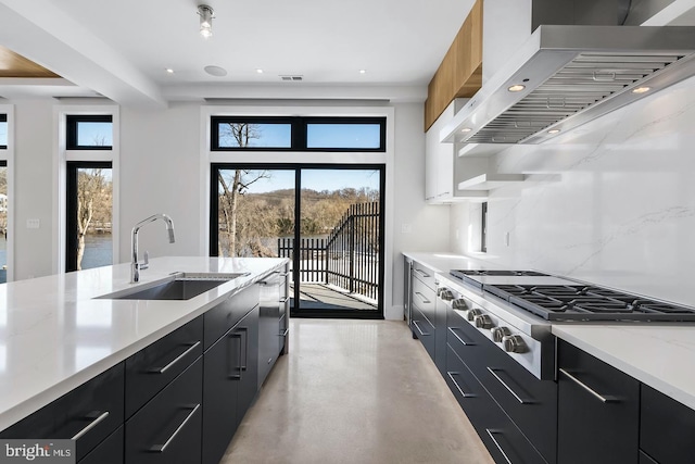 kitchen featuring light stone countertops, backsplash, stainless steel cooktop, wall chimney range hood, and sink