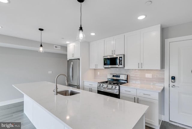 kitchen featuring a kitchen island with sink, sink, appliances with stainless steel finishes, decorative light fixtures, and white cabinetry