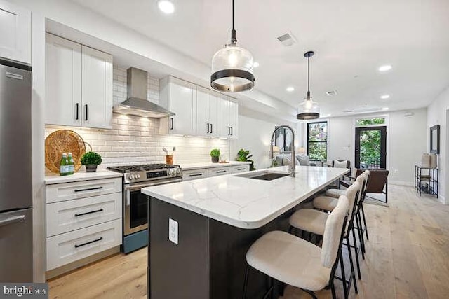 kitchen with wall chimney exhaust hood, stainless steel appliances, sink, white cabinetry, and an island with sink