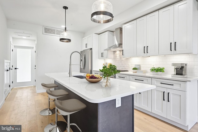 kitchen featuring wall chimney range hood, stainless steel fridge, an island with sink, decorative light fixtures, and white cabinets