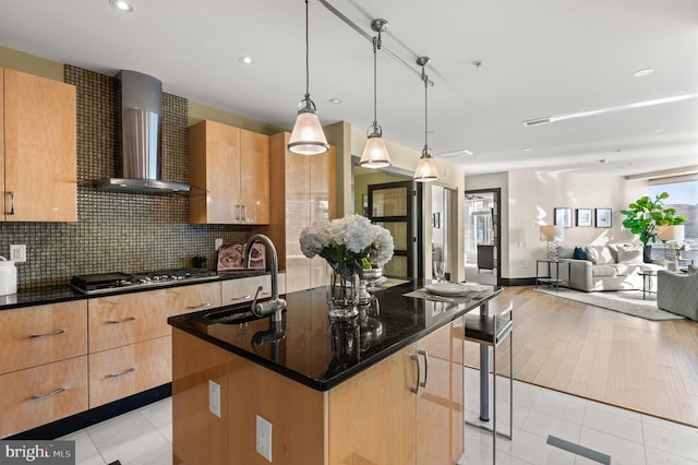 kitchen with sink, wall chimney exhaust hood, an island with sink, decorative light fixtures, and stainless steel gas cooktop