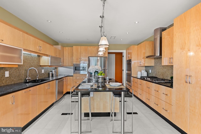 kitchen featuring appliances with stainless steel finishes, wall chimney exhaust hood, sink, a center island with sink, and decorative light fixtures