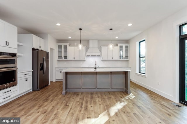 kitchen featuring appliances with stainless steel finishes, custom range hood, a kitchen island with sink, white cabinets, and hanging light fixtures