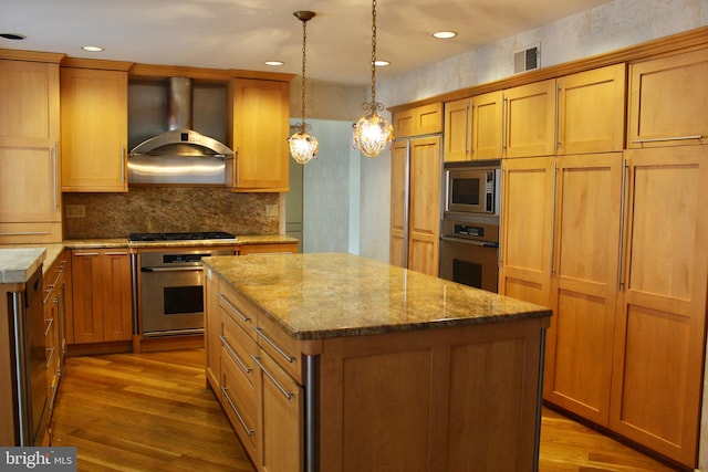 kitchen featuring appliances with stainless steel finishes, hanging light fixtures, wood-type flooring, a kitchen island, and wall chimney exhaust hood