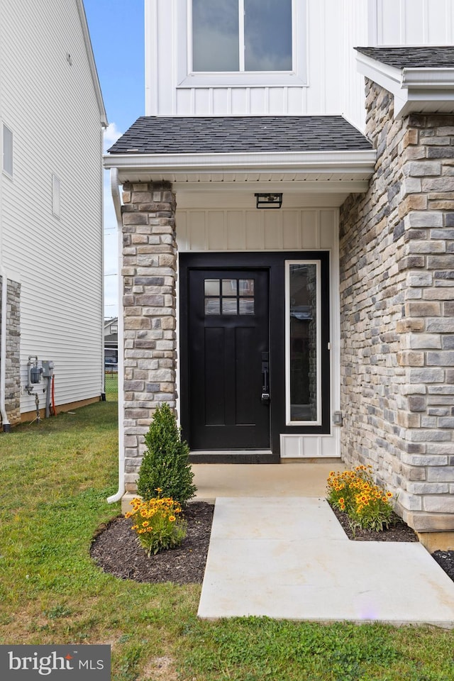 entrance to property featuring a yard, stone siding, roof with shingles, and board and batten siding