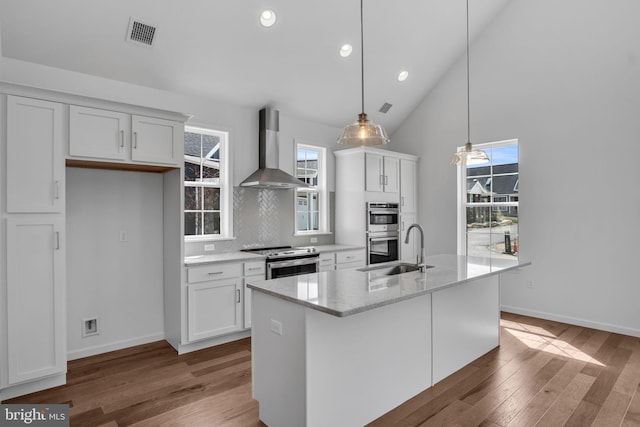 kitchen with wall chimney range hood, decorative light fixtures, a kitchen island with sink, white cabinets, and appliances with stainless steel finishes