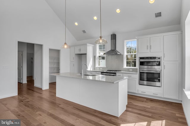 kitchen with white cabinetry, sink, wall chimney exhaust hood, decorative light fixtures, and a center island with sink