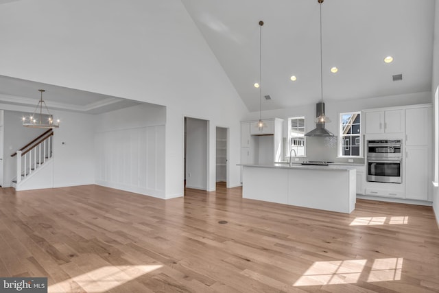 interior space featuring a center island with sink, white cabinets, wall chimney exhaust hood, double oven, and decorative light fixtures