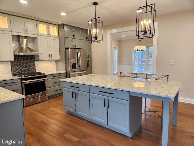 kitchen featuring hardwood / wood-style floors, appliances with stainless steel finishes, wall chimney exhaust hood, decorative backsplash, and decorative light fixtures