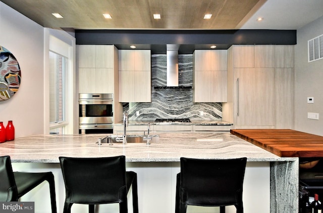 kitchen with light stone countertops, sink, wall chimney range hood, a kitchen bar, and appliances with stainless steel finishes