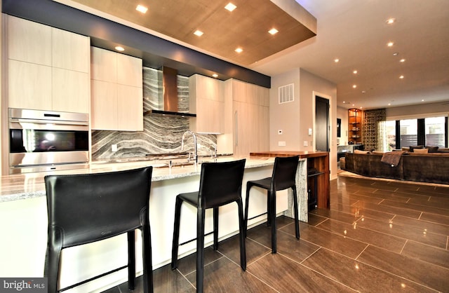 kitchen featuring light stone countertops, sink, wall chimney range hood, a kitchen breakfast bar, and double oven