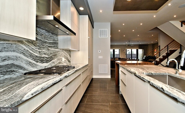 kitchen with sink, wall chimney exhaust hood, light stone counters, stainless steel gas stovetop, and a tray ceiling