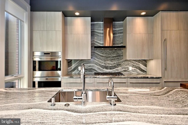 kitchen with wall chimney exhaust hood, gas stovetop, light stone countertops, tasteful backsplash, and stainless steel double oven