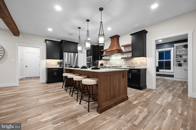 kitchen with decorative backsplash, premium range hood, a kitchen island with sink, and decorative light fixtures