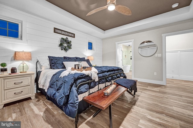 bedroom with light wood-type flooring, ensuite bathroom, a raised ceiling, and ceiling fan