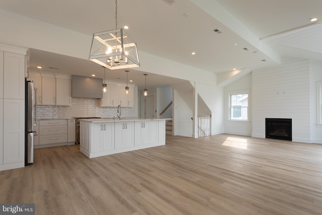 kitchen with wall chimney range hood, vaulted ceiling with beams, decorative light fixtures, a center island with sink, and appliances with stainless steel finishes