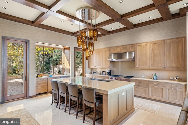 kitchen with coffered ceiling, light tile floors, a kitchen island with sink, wall chimney range hood, and hanging light fixtures