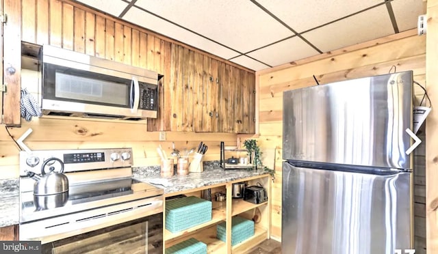 kitchen with a paneled ceiling, wood walls, appliances with stainless steel finishes, and light stone counters