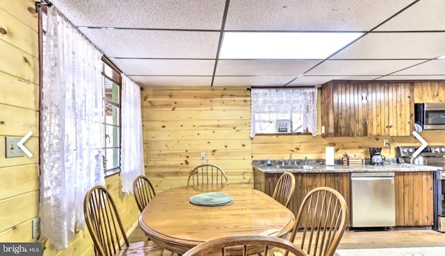 kitchen with wooden walls, appliances with stainless steel finishes, a healthy amount of sunlight, and a drop ceiling