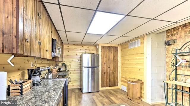 kitchen featuring appliances with stainless steel finishes, a drop ceiling, wood walls, dark stone countertops, and light hardwood / wood-style flooring