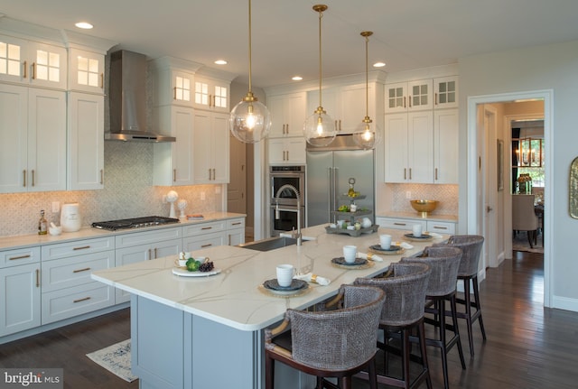 kitchen with wall chimney range hood, hanging light fixtures, stainless steel appliances, white cabinets, and a center island with sink