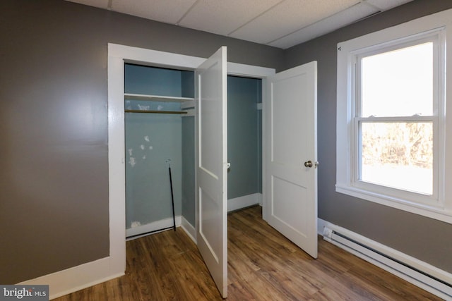 unfurnished bedroom featuring a drop ceiling, a closet, a baseboard heating unit, and dark wood-type flooring