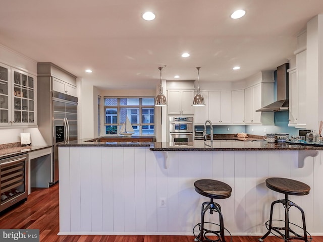 kitchen featuring wall chimney range hood, beverage cooler, appliances with stainless steel finishes, dark stone countertops, and dark hardwood / wood-style flooring