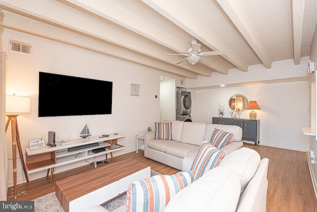 living room featuring ceiling fan, stacked washer / drying machine, light hardwood / wood-style floors, beam ceiling, and a textured ceiling