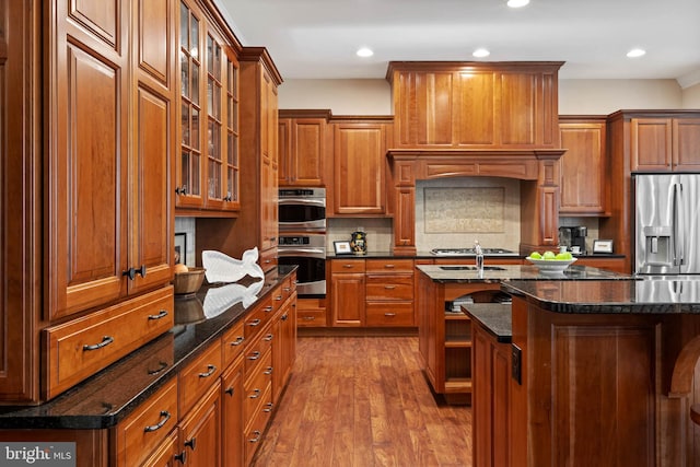 kitchen featuring dark stone counters, an island with sink, stainless steel appliances, and wood-type flooring