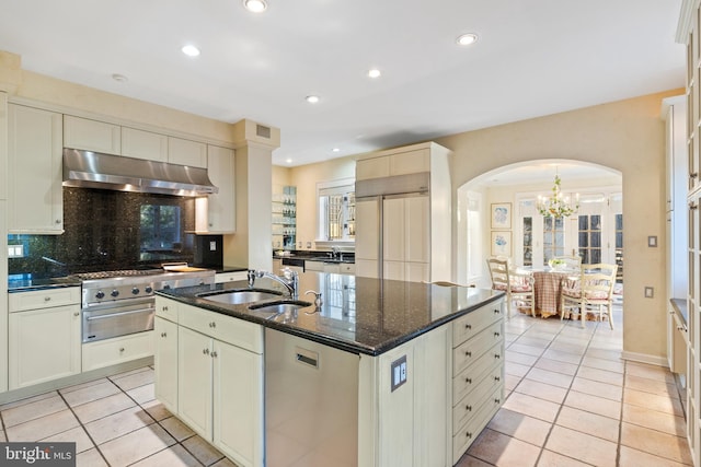 kitchen featuring wall chimney range hood, an inviting chandelier, a kitchen island with sink, and light tile floors