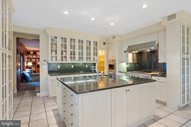 kitchen with backsplash, light tile floors, oven, a kitchen island with sink, and wall chimney exhaust hood