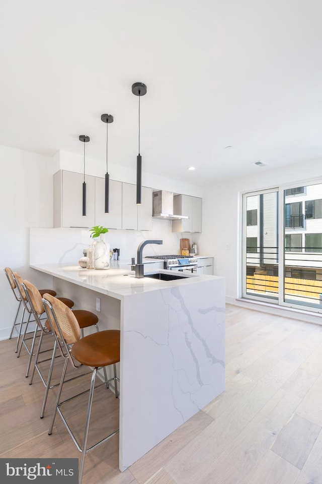 kitchen with gray cabinetry, light hardwood / wood-style flooring, wall chimney exhaust hood, hanging light fixtures, and a kitchen breakfast bar