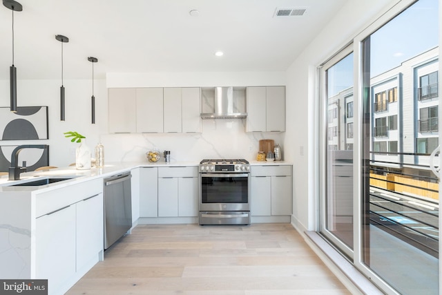 kitchen featuring hanging light fixtures, appliances with stainless steel finishes, light hardwood / wood-style floors, sink, and wall chimney exhaust hood