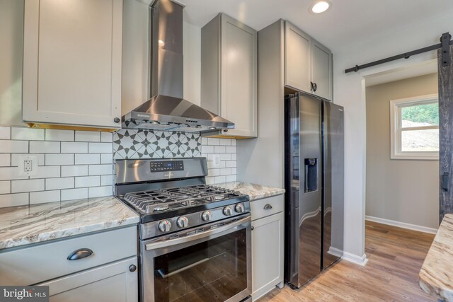 kitchen featuring light stone countertops, appliances with stainless steel finishes, light wood-type flooring, tasteful backsplash, and wall chimney exhaust hood