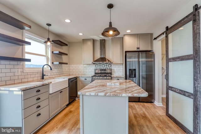 kitchen featuring gray cabinetry, a barn door, stainless steel appliances, light wood-type flooring, and wall chimney range hood