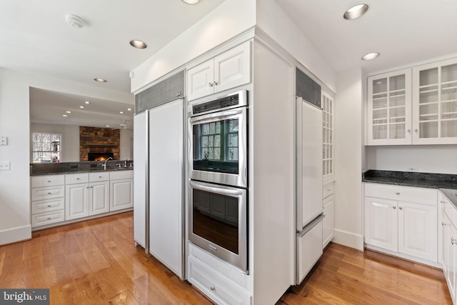 kitchen with paneled refrigerator, stainless steel double oven, light hardwood / wood-style flooring, white cabinetry, and a stone fireplace